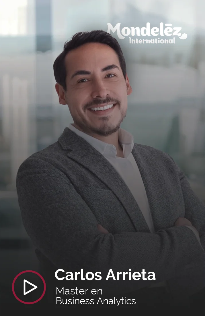 Carlos Arrieta, alumno del Master en Business Analytics, con una sonrisa confiada y los brazos cruzados, frente a la cámara durante una entrevista para Esden Business School. Carlos viste un blazer gris oscuro y una camisa blanca, proyectando una imagen profesional y segura. El logotipo de Mondelēz International se encuentra en la parte superior derecha de la imagen, indicando su afiliación con la empresa. De fondo, se distingue un entorno de oficina moderna con paredes de cristal que dejan pasar luz natural.