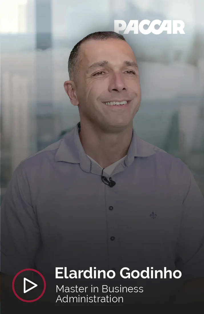 Elardino Godinho, alumno del Master in Business Administration, sonriendo durante una entrevista. Viste una camisa gris claro con un micrófono de solapa y al fondo se observa un ambiente de oficina iluminado con luz natural. El logotipo de PACCAR se muestra en la esquina superior derecha.